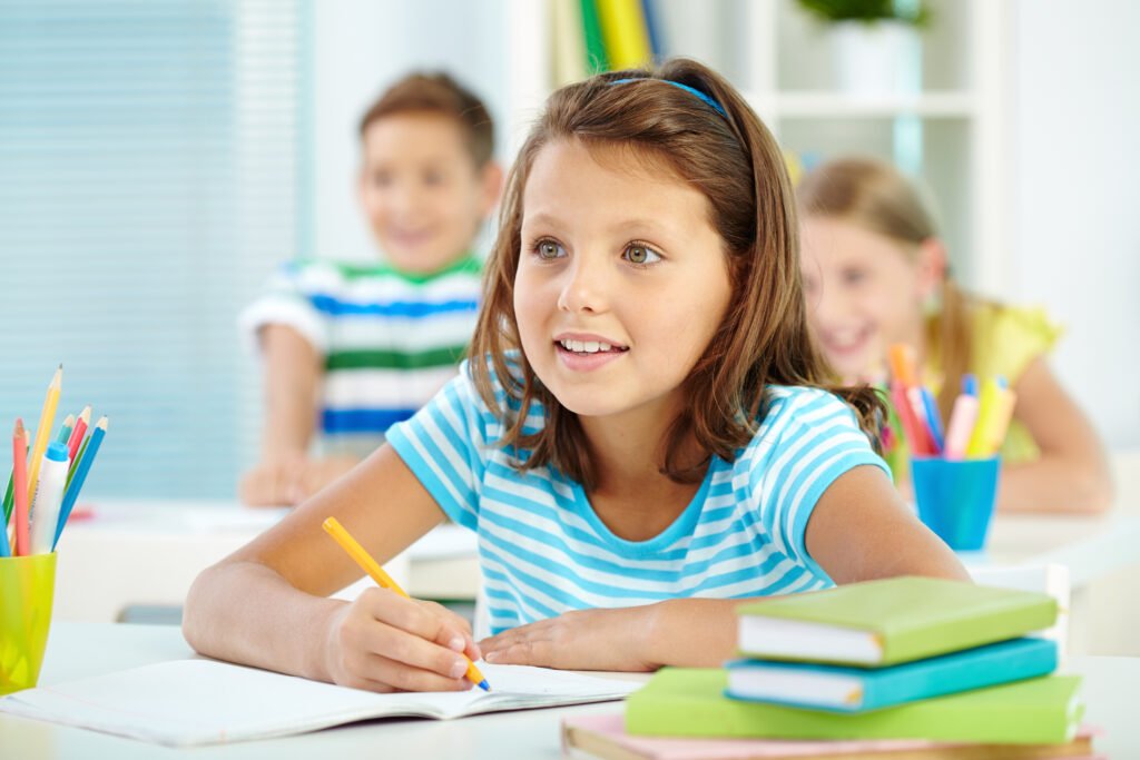 Grundschülerin schreibt aufmerksam in ihr Heft, im Hintergrund lernen zwei weitere Kinder