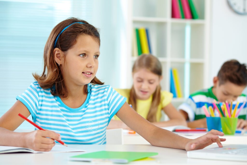 Grundschülerin hält einen Stift und überlegt bei einer Aufgabe, im Hintergrund arbeiten weitere Kinder