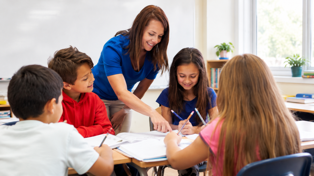 Lehrkraft beugt sich über einen Tisch und erklärt zwei Kindern eine Aufgabe